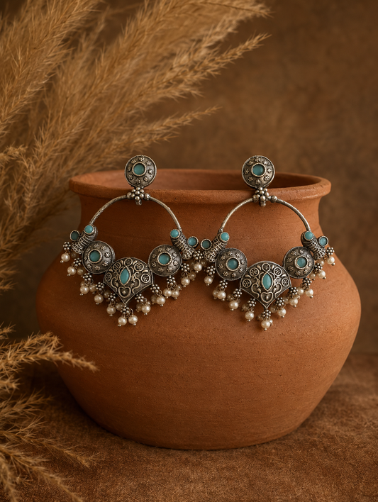 A pair of large Bhoomi oxidized silver statement hoop earrings featuring traditional tribal crescent details inlaid with light turquoise gemstones and finished with a fringe of tiny hanging pearls, draped on a terracotta pot.