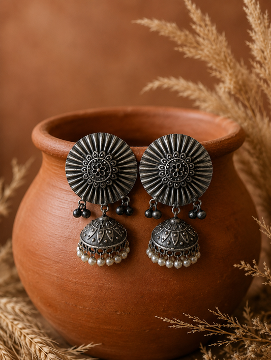 A pair of Isha oxidized silver jhumka earrings featuring large, circular sunburst studs with a floral center design and dangling dome-shaped bells trimmed with white pearls, displayed on a terracotta pot.