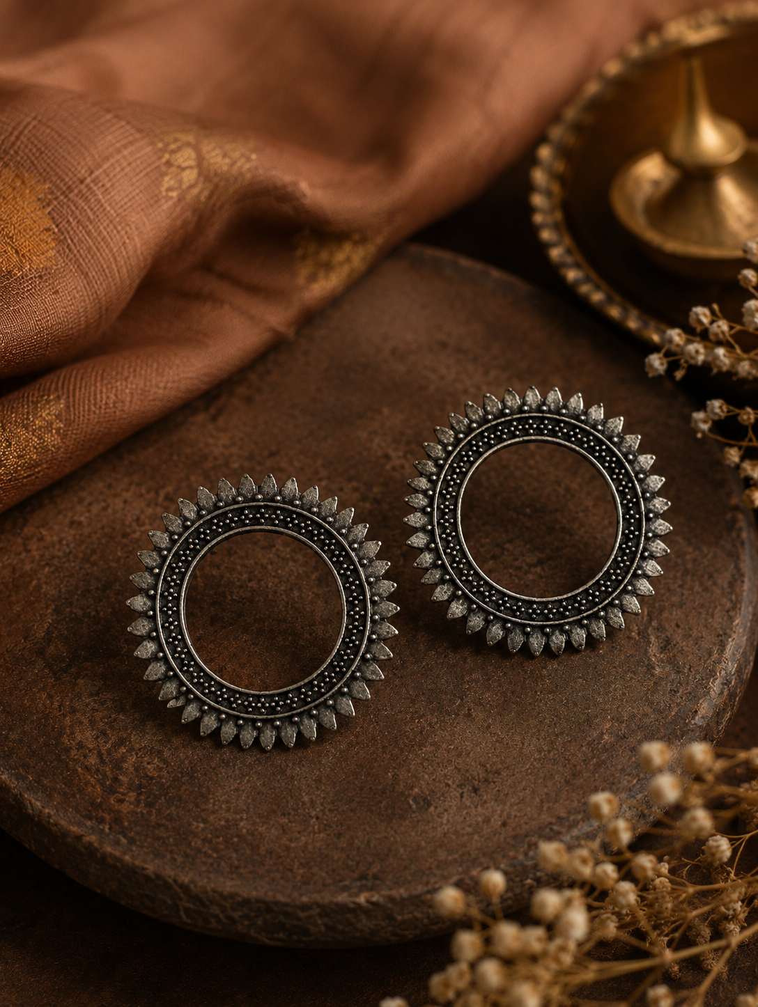 A pair of Piyusha oxidized silver open circle stud earrings with a detailed sunburst or textured petal-style outer border, displayed on a rustic wooden block.