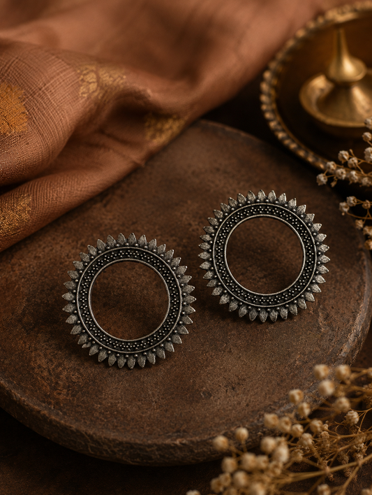 A pair of Piyusha oxidized silver open circle stud earrings with a detailed sunburst or textured petal-style outer border, displayed on a rustic wooden block.