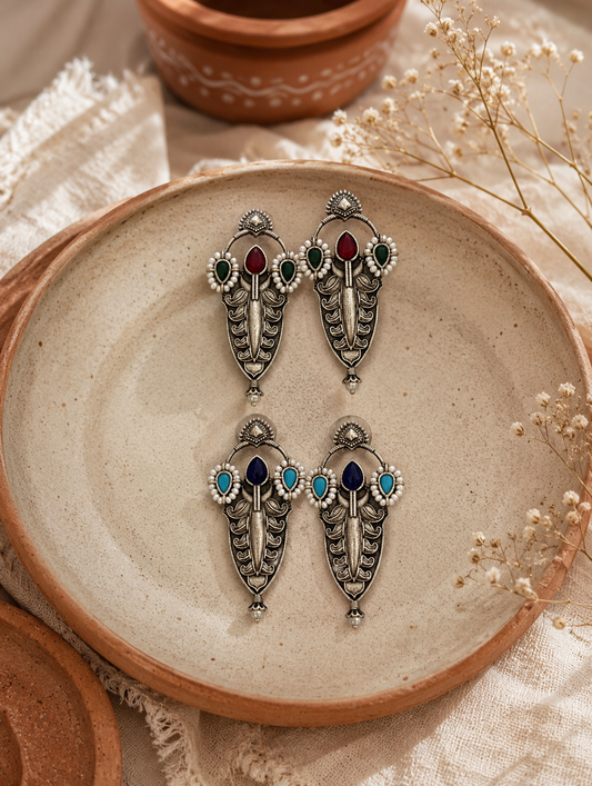 Two pairs of long oxidized silver shield earrings named Siri, decorated with colorful teardrop stones in red, green, dark blue, and light blue, lined with small white pearls inside a beige ceramic plate 