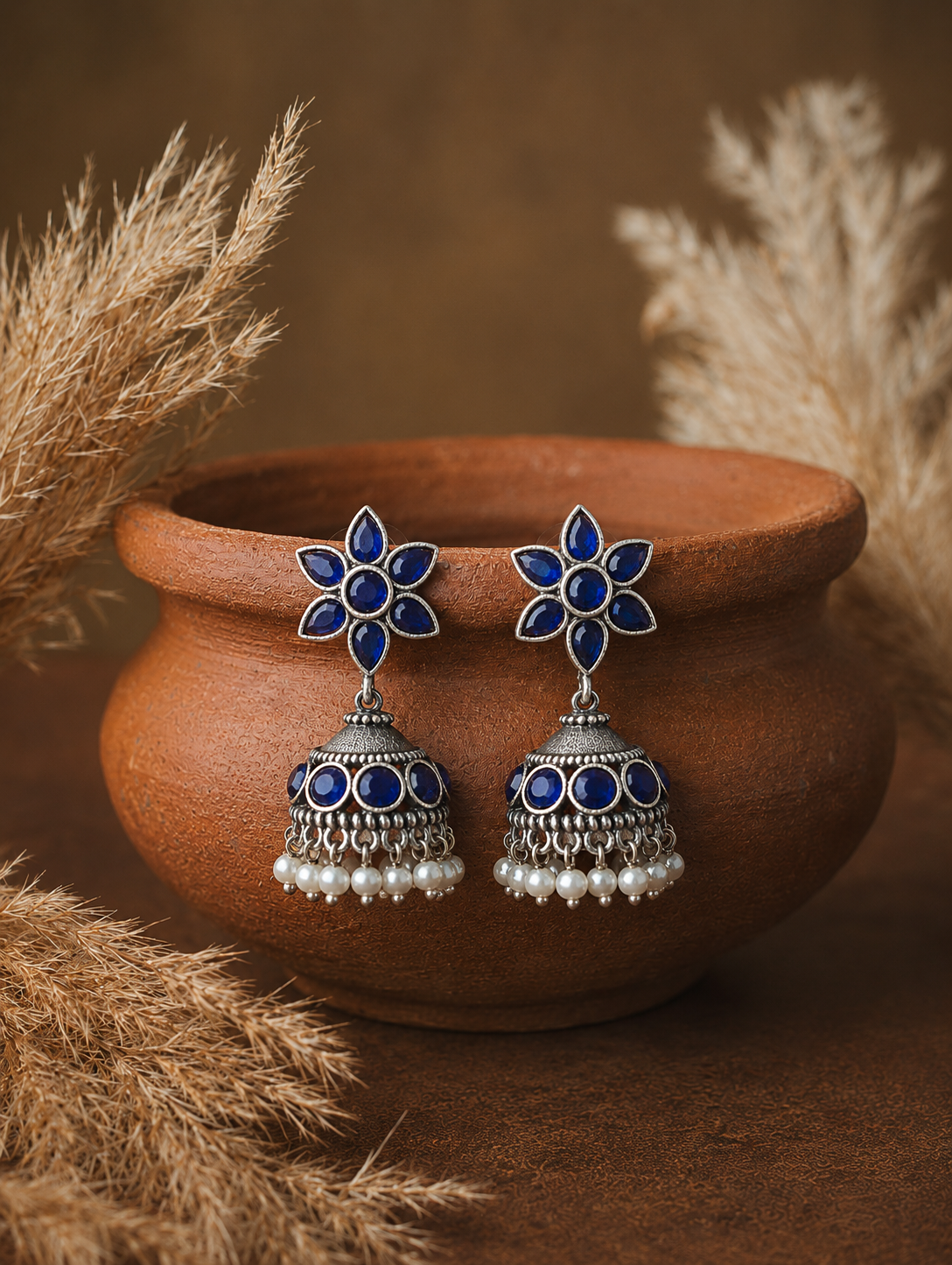 A pair of oxidized silver Vilma Jhumka earrings showcasing six-petal blue gemstone flower studs, matching blue stone circles on the domes, and hanging white pearl beads, balanced on the rim of a clay pot