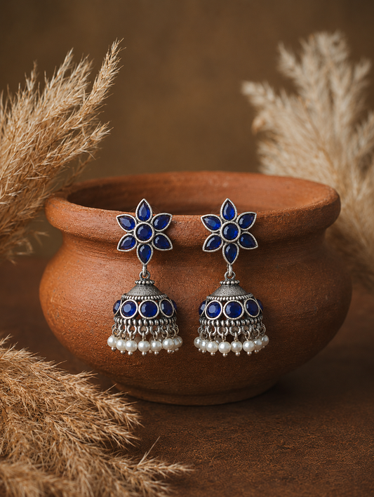 A pair of oxidized silver Vilma Jhumka earrings showcasing six-petal blue gemstone flower studs, matching blue stone circles on the domes, and hanging white pearl beads, balanced on the rim of a clay pot