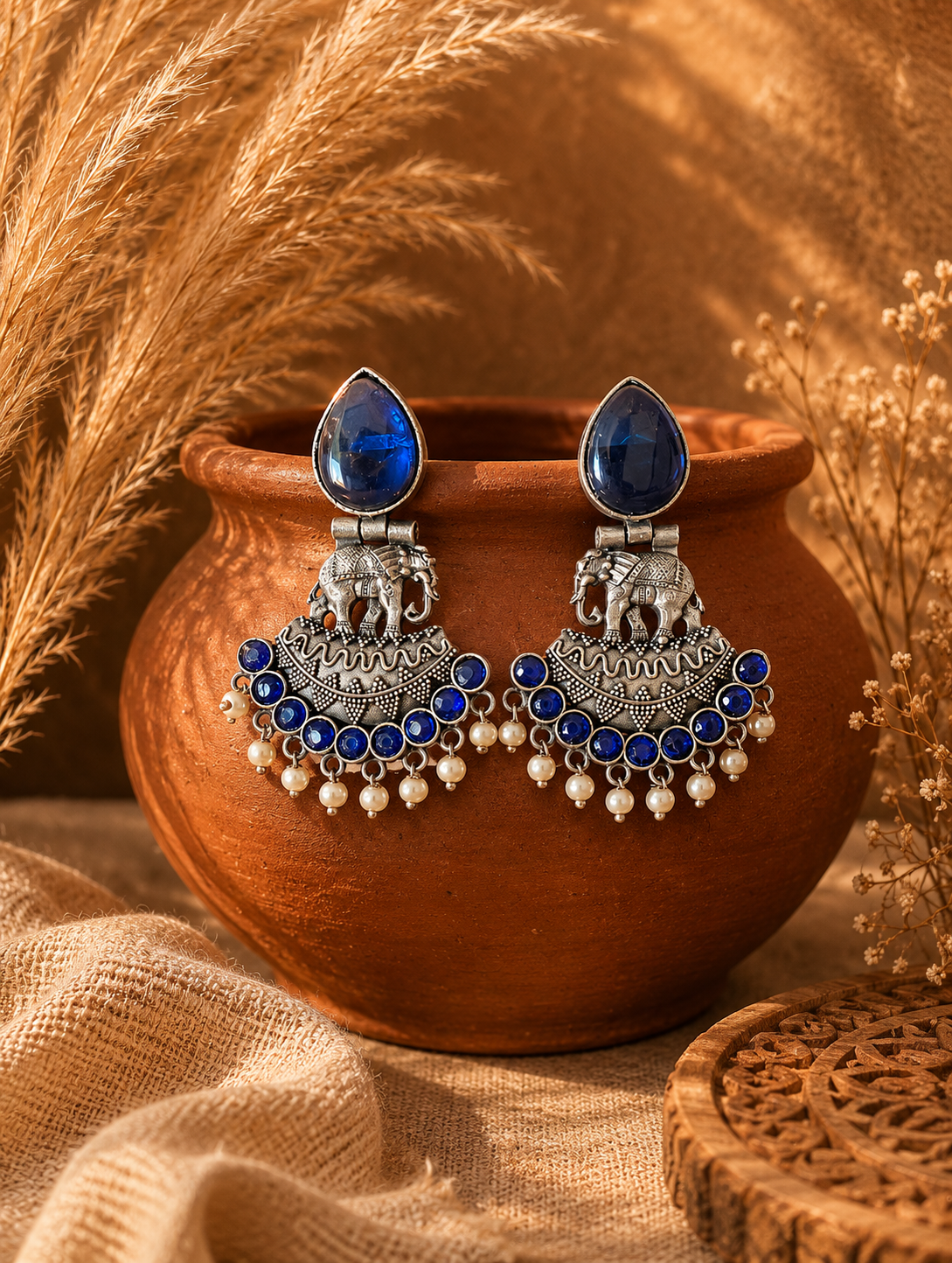 A pair of oxidized silver Nirma chandbali earrings featuring large blue teardrop stone studs, detailed elephant motifs, and a crescent base lined with small blue stones and dangling white pearls, draped on a terracotta pot 