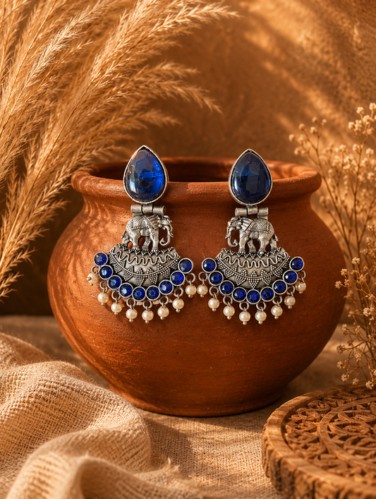 A pair of oxidized silver Nirma chandbali earrings featuring large blue teardrop stone studs, detailed elephant motifs, and a crescent base lined with small blue stones and dangling white pearls, draped on a terracotta pot 