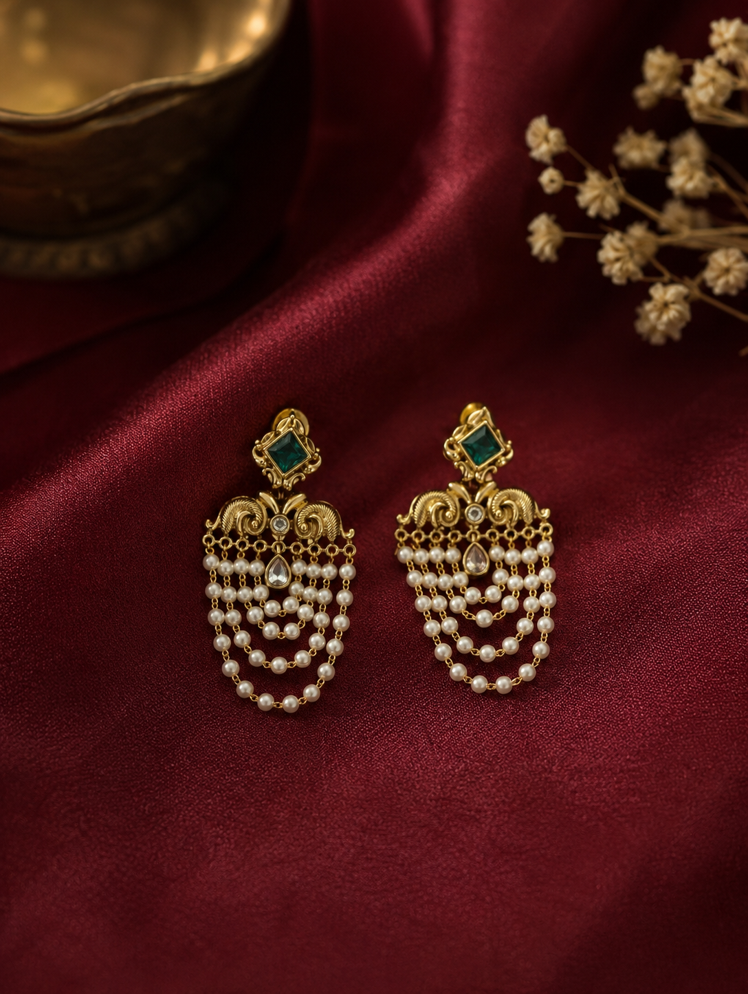 Infuse your festive wardrobe with regal elegance with these stunning traditional chandelier earrings. Designed to capture the grandeur of heritage jewelry, each earring features a striking square-cut focal stone—available in a deep emerald green or a classic clear crystal—framed in an ornate gold-plated setting. Below the main stud, intricate golden filigree work resembling stylized peacock motifs gives way to multiple cascading tiers of delicate, luminous seed pearls. These fluid, layered pearl chains crea