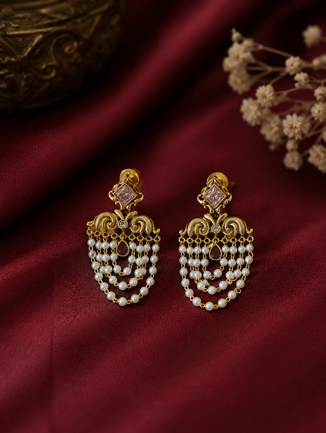 Infuse your festive wardrobe with regal elegance with these stunning traditional chandelier earrings. Designed to capture the grandeur of heritage jewelry, each earring features a striking square-cut focal stone—available in a deep emerald green or a classic clear crystal—framed in an ornate gold-plated setting. Below the main stud, intricate golden filigree work resembling stylized peacock motifs gives way to multiple cascading tiers of delicate, luminous seed pearls. These fluid, layered pearl chains crea