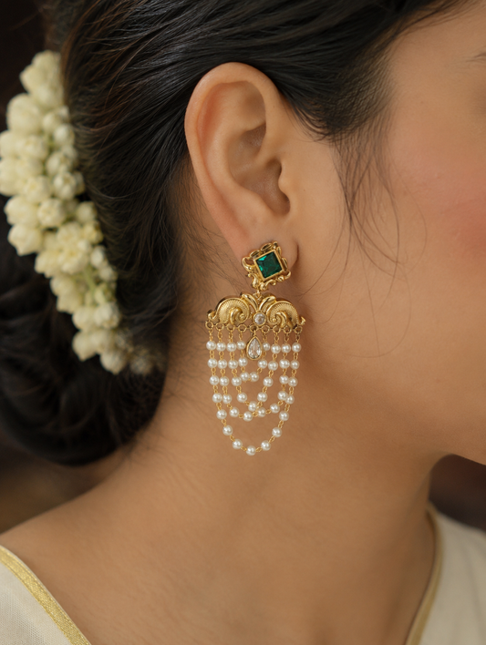 Infuse your festive wardrobe with regal elegance with these stunning traditional chandelier earrings. Designed to capture the grandeur of heritage jewelry, each earring features a striking square-cut focal stone—available in a deep emerald green or a classic clear crystal—framed in an ornate gold-plated setting. Below the main stud, intricate golden filigree work resembling stylized peacock motifs gives way to multiple cascading tiers of delicate, luminous seed pearls. These fluid, layered pearl chains crea