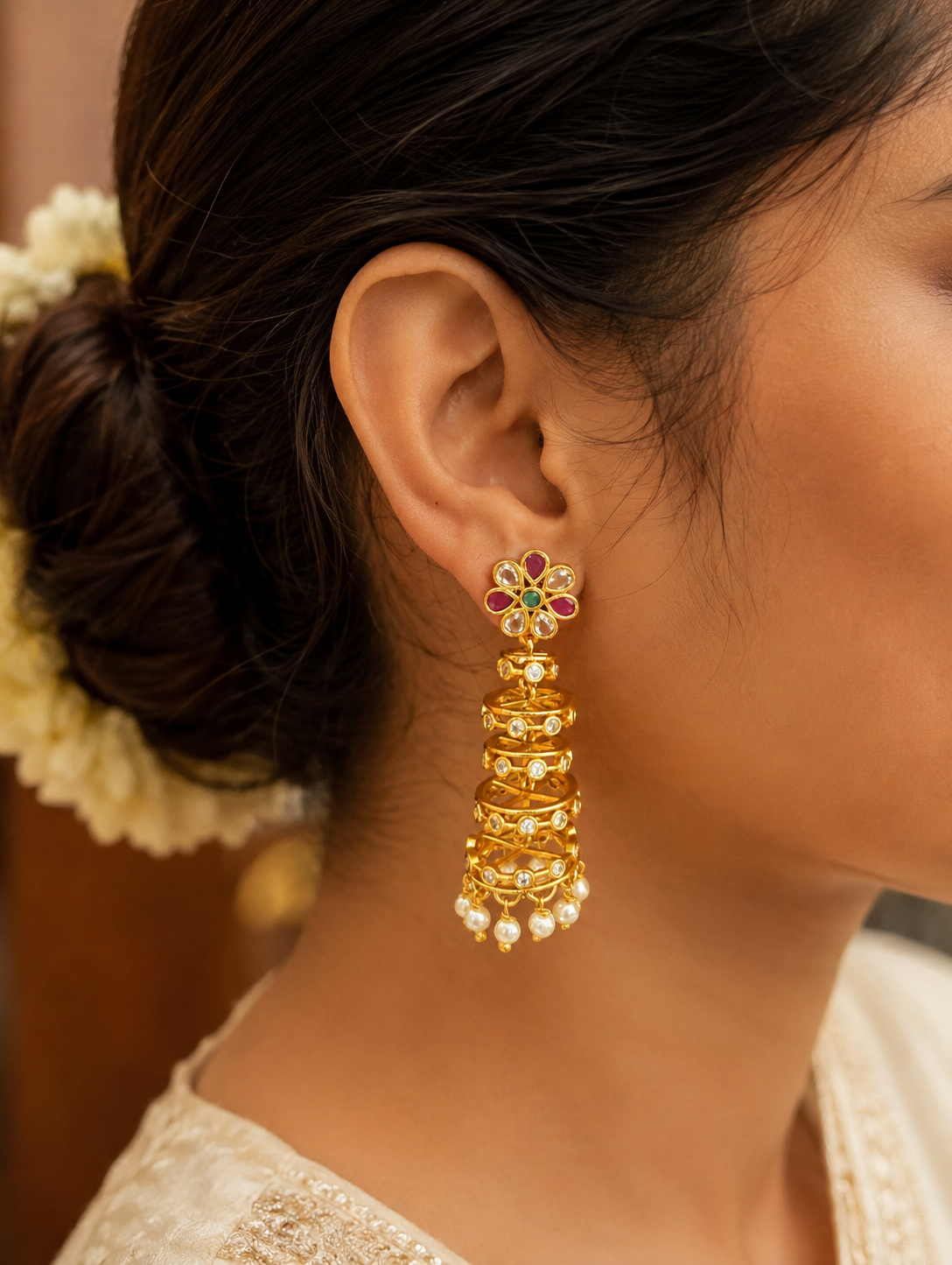 Make a magnificent style statement with these exquisite multi-tiered jhumka earrings, masterfully blending classic ethnic charm with a striking contemporary frame. The earrings feature a vibrant, flower-shaped stud accented with alternating ruby-pink and pristine clear Kundan-style stones, anchored by a rich green central gem. Suspending from the floral cluster is a dramatic, architectural cascade of four gold-plated concentric cages, each step meticulously detailed with delicate openwork and inlaid circula