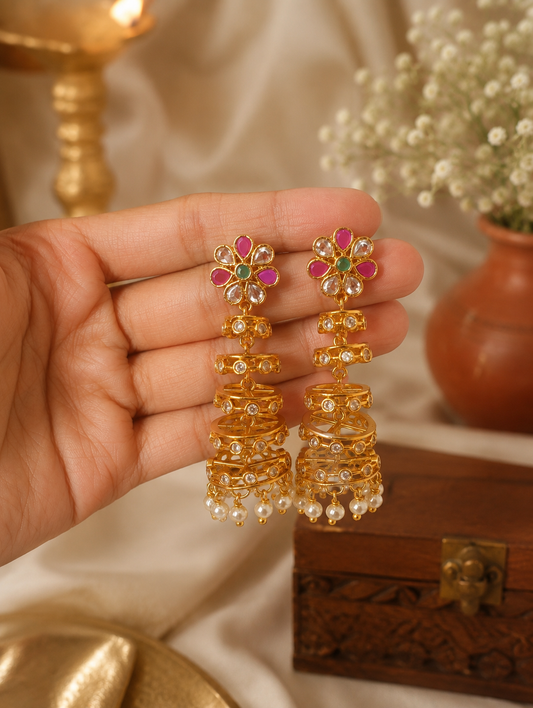 Make a magnificent style statement with these exquisite multi-tiered jhumka earrings, masterfully blending classic ethnic charm with a striking contemporary frame. The earrings feature a vibrant, flower-shaped stud accented with alternating ruby-pink and pristine clear Kundan-style stones, anchored by a rich green central gem. Suspending from the floral cluster is a dramatic, architectural cascade of four gold-plated concentric cages, each step meticulously detailed with delicate openwork and inlaid circula