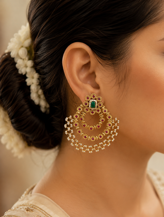 Channel timeless grandeur with these magnificent multi-tiered Chandbali earrings, beautifully designed to celebrate heritage artistry. Featuring a classic crescent silhouette, each earring begins with an ornate floral-framed stud centered around a striking, rectangular emerald-green gemstone. Suspended below are three concentric, cascading crescent tracks adorned with vibrant ruby-pink accents set against a rich matte gold finish. The outermost tier boasts an exquisite, openwork lattice framework studded wi
