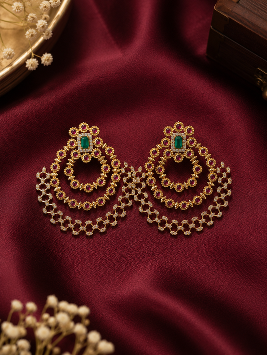 Channel timeless grandeur with these magnificent multi-tiered Chandbali earrings, beautifully designed to celebrate heritage artistry. Featuring a classic crescent silhouette, each earring begins with an ornate floral-framed stud centered around a striking, rectangular emerald-green gemstone. Suspended below are three concentric, cascading crescent tracks adorned with vibrant ruby-pink accents set against a rich matte gold finish. The outermost tier boasts an exquisite, openwork lattice framework studded wi