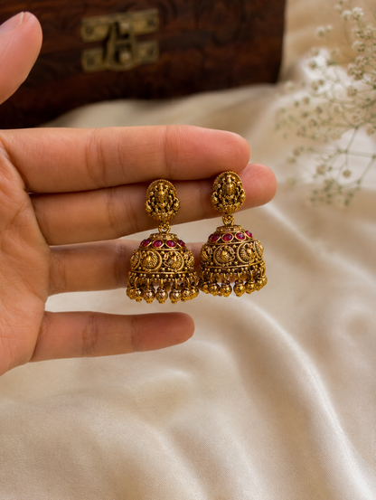 Embrace the divine essence of South Indian heritage with these meticulously crafted temple-style jhumka earrings. Designed to replicate the timeless opulence of antique jewelry, each earring features a small oval stud intricately sculpted with a traditional deity motif. The classic bell-shaped dome (jhumka) gracefully suspends below, detailed with a striking row of rich, cabochon ruby-pink stones along the top rim and ornate filigree patterns depicting classic peacock accents throughout the center. Finished