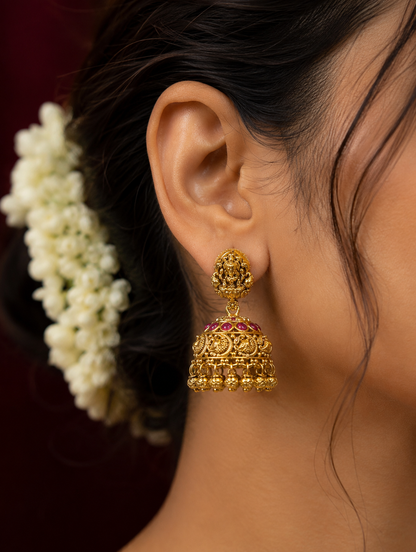 Embrace the divine essence of South Indian heritage with these meticulously crafted temple-style jhumka earrings. Designed to replicate the timeless opulence of antique jewelry, each earring features a small oval stud intricately sculpted with a traditional deity motif. The classic bell-shaped dome (jhumka) gracefully suspends below, detailed with a striking row of rich, cabochon ruby-pink stones along the top rim and ornate filigree patterns depicting classic peacock accents throughout the center. Finished