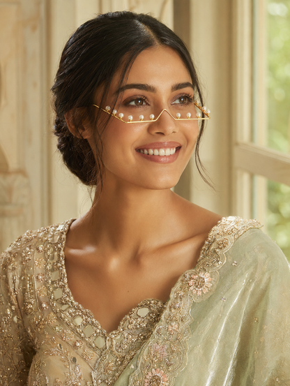 Minimalist gold wire facial frame accessory shaped like glasses, studded with luminous pearls along the lower rim and temples, displayed on a soft beige fabric background and worn by a smiling model in traditional attire.