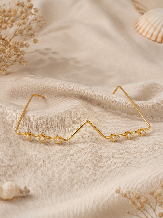 Minimalist gold wire facelet accessory designed like a pair of glasses, decorated with small golden flower motifs and central white pearls along the bottom rim, shown on a textured cream backdrop and modeled by a woman in a sage green saree.