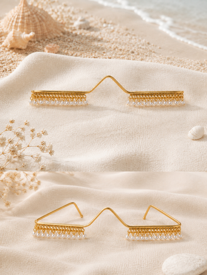 Avant-garde gold wire facial jewelry frame shaped like glasses, featuring a delicate fringe of hanging white pearls along the lower rim, displayed against a beach-themed setup and worn by a smiling model in an embroidered mint green saree.