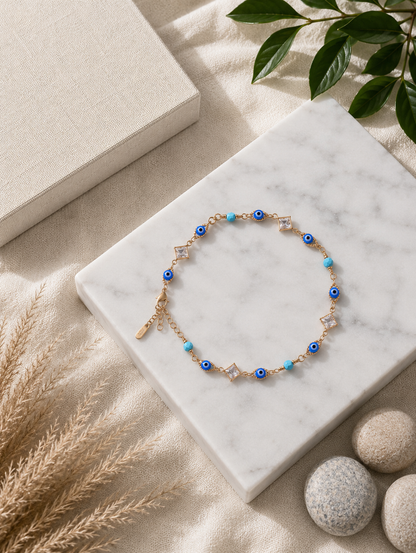 A top-down product shot of a gold-toned chain anklet arranged in a circle on a square white marble slab. The anklet features alternating round blue and white Evil Eye charms, bright turquoise beads, and clear square-cut crystals in diamond-shaped gold frames. The background has an earthy, beach-inspired aesthetic with textured fabric, dried grass, smooth river stones, and green leaves.