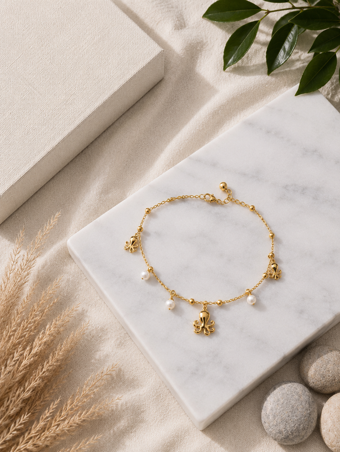 A close-up lifestyle photograph of a model's ankle adorned with a gold-toned charm anklet. The delicate chain features a collection of dangling nautical charms, including a central octopus, an anchor, a small seashell, and petite white pearls. The model is wearing light blue denim, set against a bright, warm-toned background with minimalist pottery, smooth stones, and dried stalks.