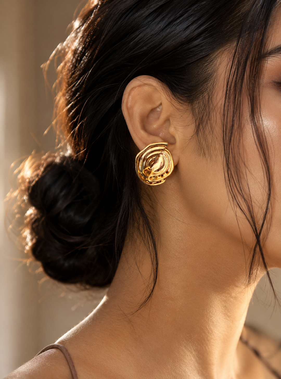 A side-profile lifestyle photograph showing a woman with dark hair styled in a loose bun, wearing a large, sculptural gold stud earring from the Spiral collection. The earring features an organic, textured vortex design with a high-shine, molten metal finish that catches the light. As seen in GE001_spiral_599.png.