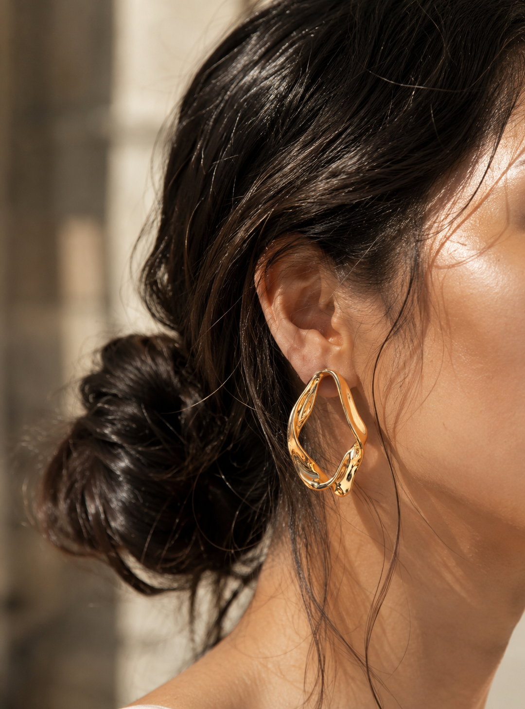 A side-profile lifestyle photograph of a woman with her dark hair swept up in a loose, textured bun, showcasing an oversized, abstract gold hoop earring from the Golden Twist collection. The earring features a thick, fluidly contorted ribbon design with a mirror-shine molten finish reflecting soft sunlight. As seen in GE002_gold twist_599.png.