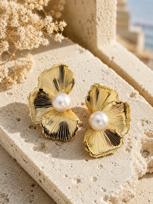 A flat-lay product photograph of a pair of oversized gold stud earrings from the Flower Pearl collection, displayed on a porous white travertine stone blocks under warm sunlight. Each earring showcases a textured, fan-shaped golden floral design with finely ribbed lines, centering a large, luminous white pearl. The background is softly styled with delicate dried white flowers. As seen in GE003_flower pearl_699.png.