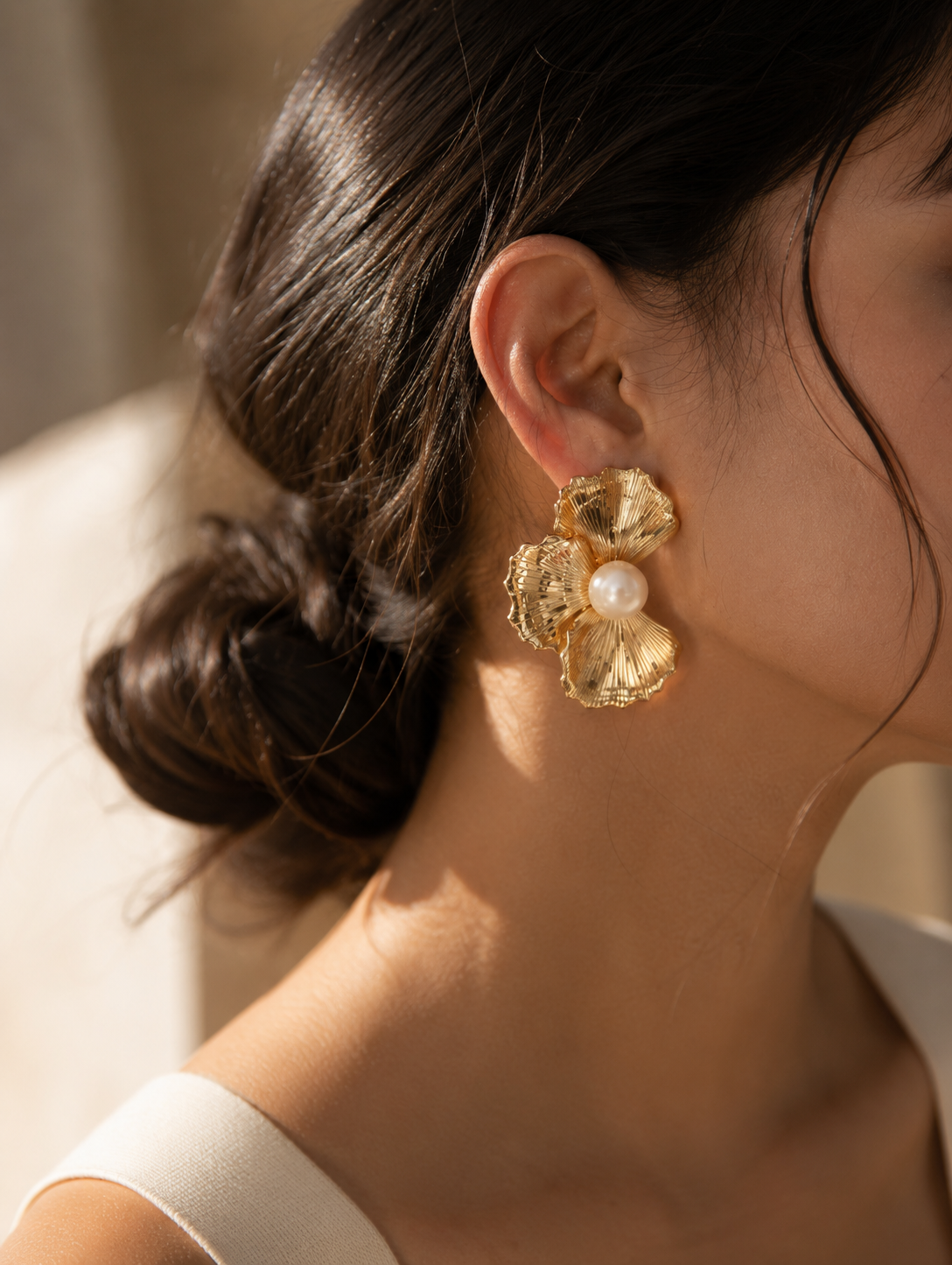 A flat-lay product photograph of a pair of oversized gold stud earrings from the Flower Pearl collection, displayed on a porous white travertine stone blocks under warm sunlight. Each earring showcases a textured, fan-shaped golden floral design with finely ribbed lines, centering a large, luminous white pearl. The background is softly styled with delicate dried white flowers. As seen in GE003_flower pearl_699.png.