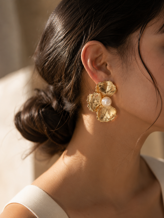 A flat-lay product photograph of a pair of oversized gold stud earrings from the Flower Pearl collection, displayed on a porous white travertine stone blocks under warm sunlight. Each earring showcases a textured, fan-shaped golden floral design with finely ribbed lines, centering a large, luminous white pearl. The background is softly styled with delicate dried white flowers. As seen in GE003_flower pearl_699.png.