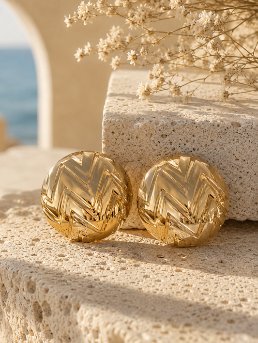 A flat-lay product photograph of a pair of large, circular gold dome stud earrings from the Semiglow collection, resting upright on textured white travertine stone under warm sunlight. The earrings feature a highly polished metallic finish with a deeply engraved geometric chevron zigzag pattern across their curved surfaces. The background shows soft, dried white flowers and an out-of-focus seaside archway view. As seen in GE004_semiglow.png.