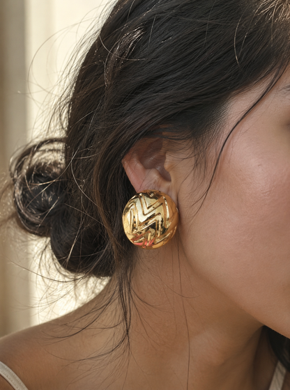A flat-lay product photograph of a pair of large, circular gold dome stud earrings from the Semiglow collection, resting upright on textured white travertine stone under warm sunlight. The earrings feature a highly polished metallic finish with a deeply engraved geometric chevron zigzag pattern across their curved surfaces. The background shows soft, dried white flowers and an out-of-focus seaside archway view. As seen in GE004_semiglow.png.