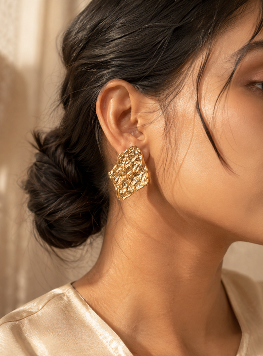 A flat-lay product photograph of a pair of large, abstract diamond-shaped gold stud earrings from the Stone Arc collection. The earrings are displayed upright on a porous cream-colored travertine stone under bright, natural sunlight, showcasing an intensely crumpled and textured molten metal finish. The background is softly styled with delicate dried white flowers. As seen in GE005_stone arc_550.png.
