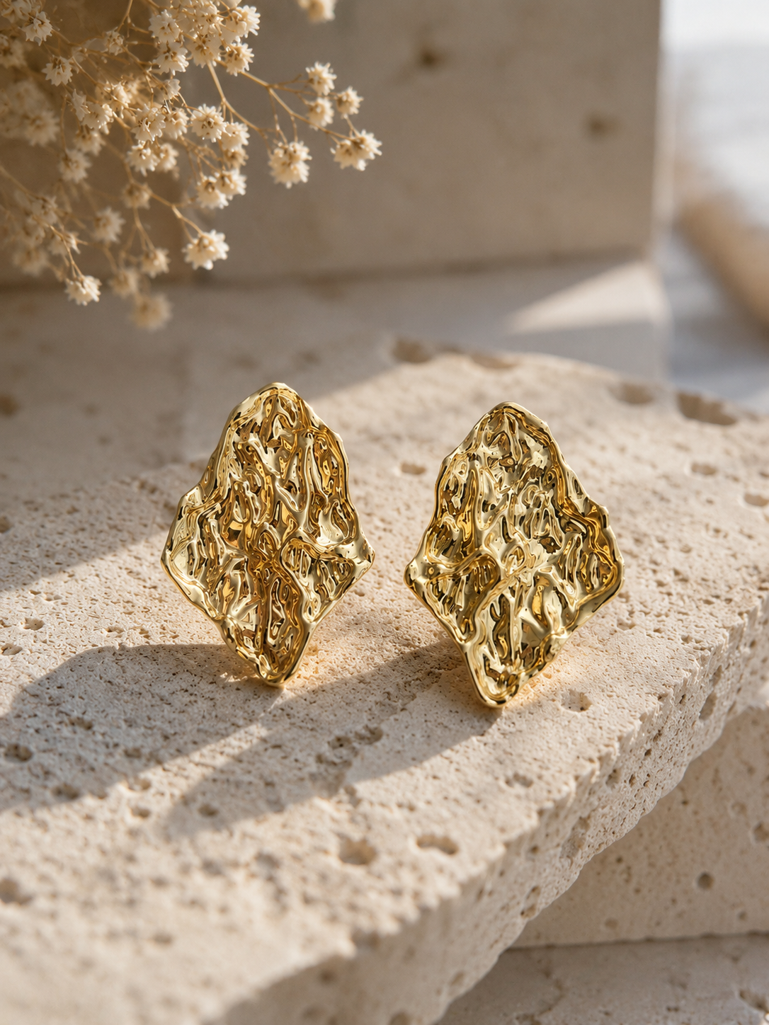 A flat-lay product photograph of a pair of large, abstract diamond-shaped gold stud earrings from the Stone Arc collection. The earrings are displayed upright on a porous cream-colored travertine stone under bright, natural sunlight, showcasing an intensely crumpled and textured molten metal finish. The background is softly styled with delicate dried white flowers. As seen in GE005_stone arc_550.png.