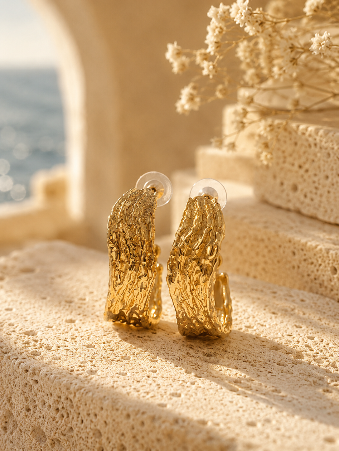 A product photograph of a pair of wide, chunky C-shaped gold hoop earrings from the Liquid Lava collection, standing upright on porous white travertine stone steps under warm sunlight. The earrings feature an intensely textured, rippled molten metal finish with clear plastic disc backings on the posts. The background is beautifully styled with dried white flowers and a blurred seaside architectural backdrop. As seen in GE006_hoops.png.