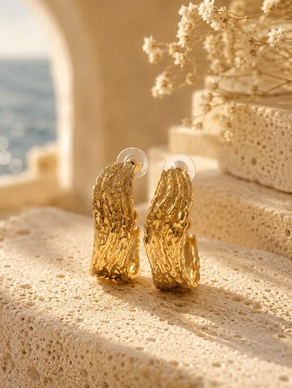 A product photograph of a pair of wide, chunky C-shaped gold hoop earrings from the Liquid Lava collection, standing upright on porous white travertine stone steps under warm sunlight. The earrings feature an intensely textured, rippled molten metal finish with clear plastic disc backings on the posts. The background is beautifully styled with dried white flowers and a blurred seaside architectural backdrop. As seen in GE006_hoops.png.