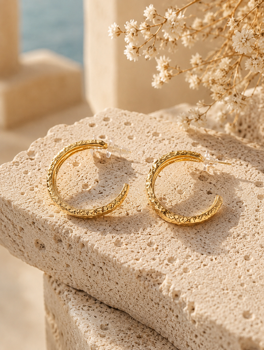 A product photograph of a pair of wide, chunky C-shaped gold hoop earrings from the Liquid Lava collection, standing upright on porous white travertine stone steps under warm sunlight. The earrings feature an intensely textured, rippled molten metal finish with clear plastic disc backings on the posts. The background is beautifully styled with dried white flowers and a blurred seaside architectural backdrop. As seen in GE006_hoops.png.