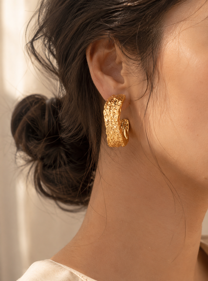 A product photograph of a pair of wide, chunky C-shaped gold hoop earrings from the Liquid Lava collection, standing upright on porous white travertine stone steps under warm sunlight. The earrings feature an intensely textured, rippled molten metal finish with clear plastic disc backings on the posts. The background is beautifully styled with dried white flowers and a blurred seaside architectural backdrop. As seen in GE006_hoops.png.