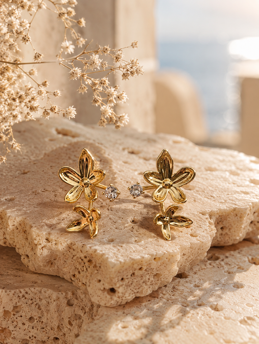 A flat-lay product photograph of a pair of gold floral ear jacket earrings from the Gemflora collection, displayed upright on a porous cream-colored travertine stone under natural sunlight. Each earring features a larger polished five-petal gold flower stud connected to a single sparkling clear crystal and a smaller dangling gold flower element below. The background is softly styled with delicate dried white baby's breath flowers and a blurred seaside backdrop. As seen in GE007_gemflora.png.