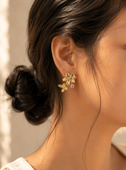 A flat-lay product photograph of a pair of gold floral ear jacket earrings from the Gemflora collection, displayed upright on a porous cream-colored travertine stone under natural sunlight. Each earring features a larger polished five-petal gold flower stud connected to a single sparkling clear crystal and a smaller dangling gold flower element below. The background is softly styled with delicate dried white baby's breath flowers and a blurred seaside backdrop. As seen in GE007_gemflora.png.