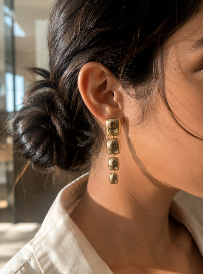 A side-profile lifestyle photograph of a woman with her dark hair swept up into a textured bun, showcasing a gold linear drop earring from the Link Chain collection. The earring features four vertically stacked, graduated, and puffy rectangular metal links with a mirror-shine polished gold finish that reflects the warm sunlight. The woman is wearing a neutral cream-colored textured linen shirt. As seen in GE008_link chain.png.