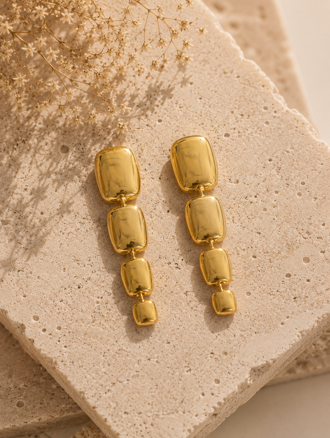 A side-profile lifestyle photograph of a woman with her dark hair swept up into a textured bun, showcasing a gold linear drop earring from the Link Chain collection. The earring features four vertically stacked, graduated, and puffy rectangular metal links with a mirror-shine polished gold finish that reflects the warm sunlight. The woman is wearing a neutral cream-colored textured linen shirt. As seen in GE008_link chain.png.