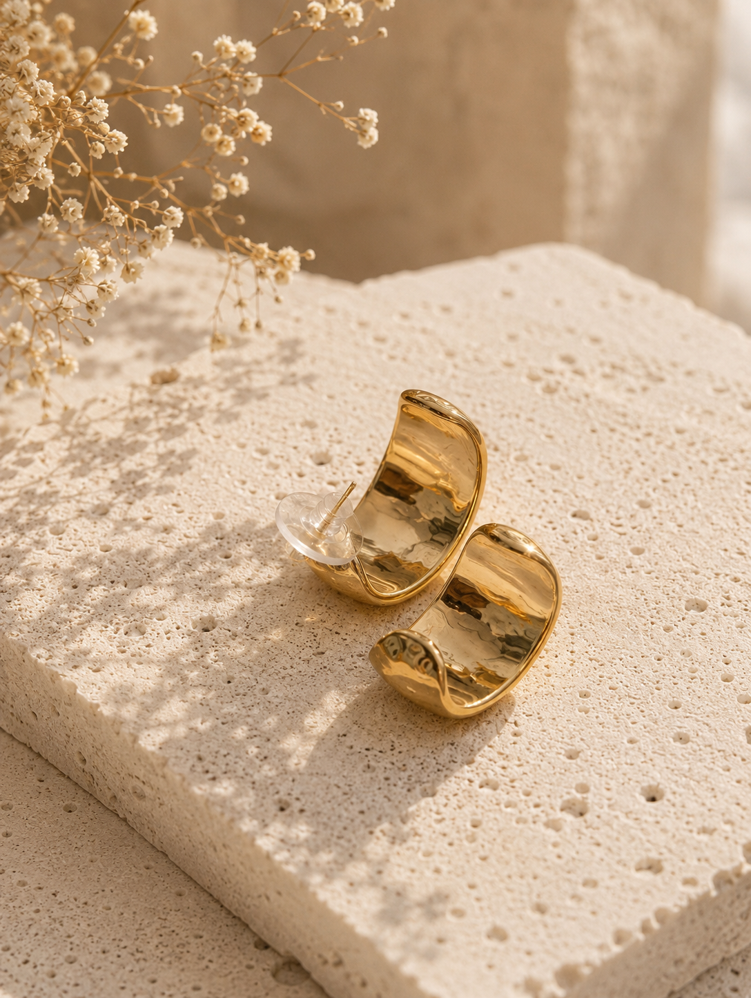A side-profile lifestyle photograph of a woman with her dark hair styled into a textured low bun, showcasing a thick gold huggie hoop earring from the Forma collection. The earring features a wide, abstractly crumpled and indented molten metal design with a high-shine mirror finish catching soft sunlight. The woman is wearing a casual cream-colored linen shirt. As seen in GE010_forma_550.png.