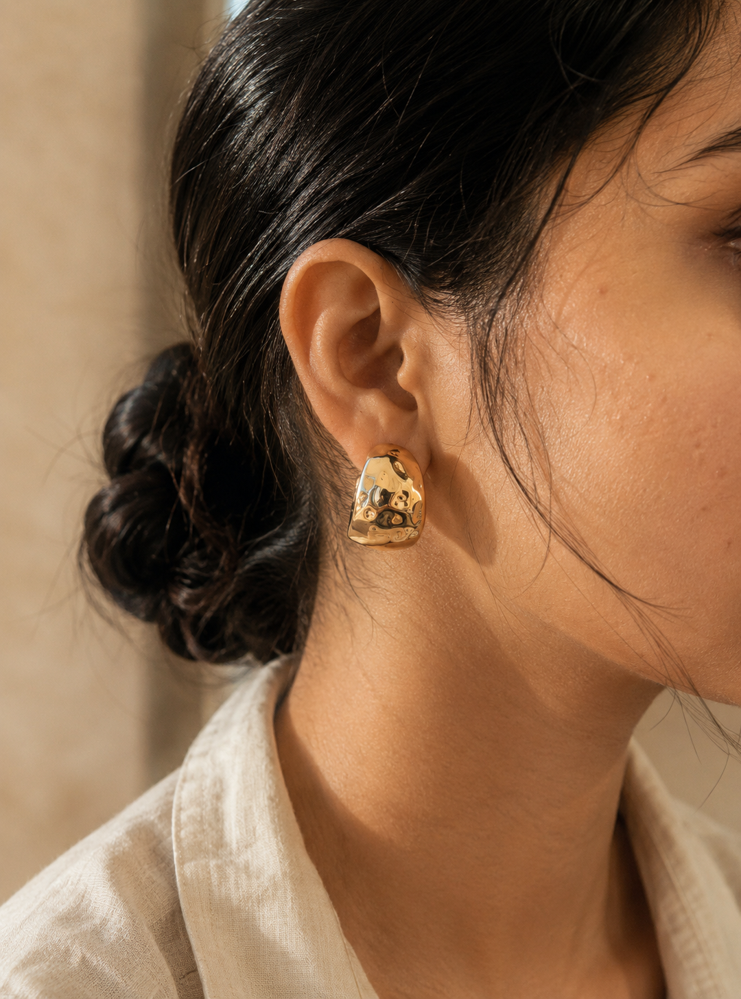 A side-profile lifestyle photograph of a woman with her dark hair styled into a textured low bun, showcasing a thick gold huggie hoop earring from the Forma collection. The earring features a wide, abstractly crumpled and indented molten metal design with a high-shine mirror finish catching soft sunlight. The woman is wearing a casual cream-colored linen shirt. As seen in GE010_forma_550.png.
