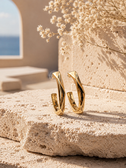 A flat-lay product photograph of a pair of oversized, elongated gold hoop earrings from the Long Twist collection, standing upright on porous white travertine stone steps under warm sunlight. The earrings feature a unique vertically stretched C-shape with an angular twisted design and a high-shine molten gold finish, fitted with clear plastic disc backings on the posts. The background is softly styled with delicate dried white flowers and a blurred Mediterranean architectural setting. As seen in GE011_long 