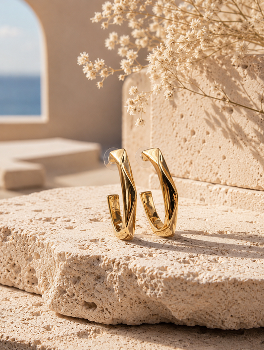 A flat-lay product photograph of a pair of oversized, elongated gold hoop earrings from the Long Twist collection, standing upright on porous white travertine stone steps under warm sunlight. The earrings feature a unique vertically stretched C-shape with an angular twisted design and a high-shine molten gold finish, fitted with clear plastic disc backings on the posts. The background is softly styled with delicate dried white flowers and a blurred Mediterranean architectural setting. As seen in GE011_long 