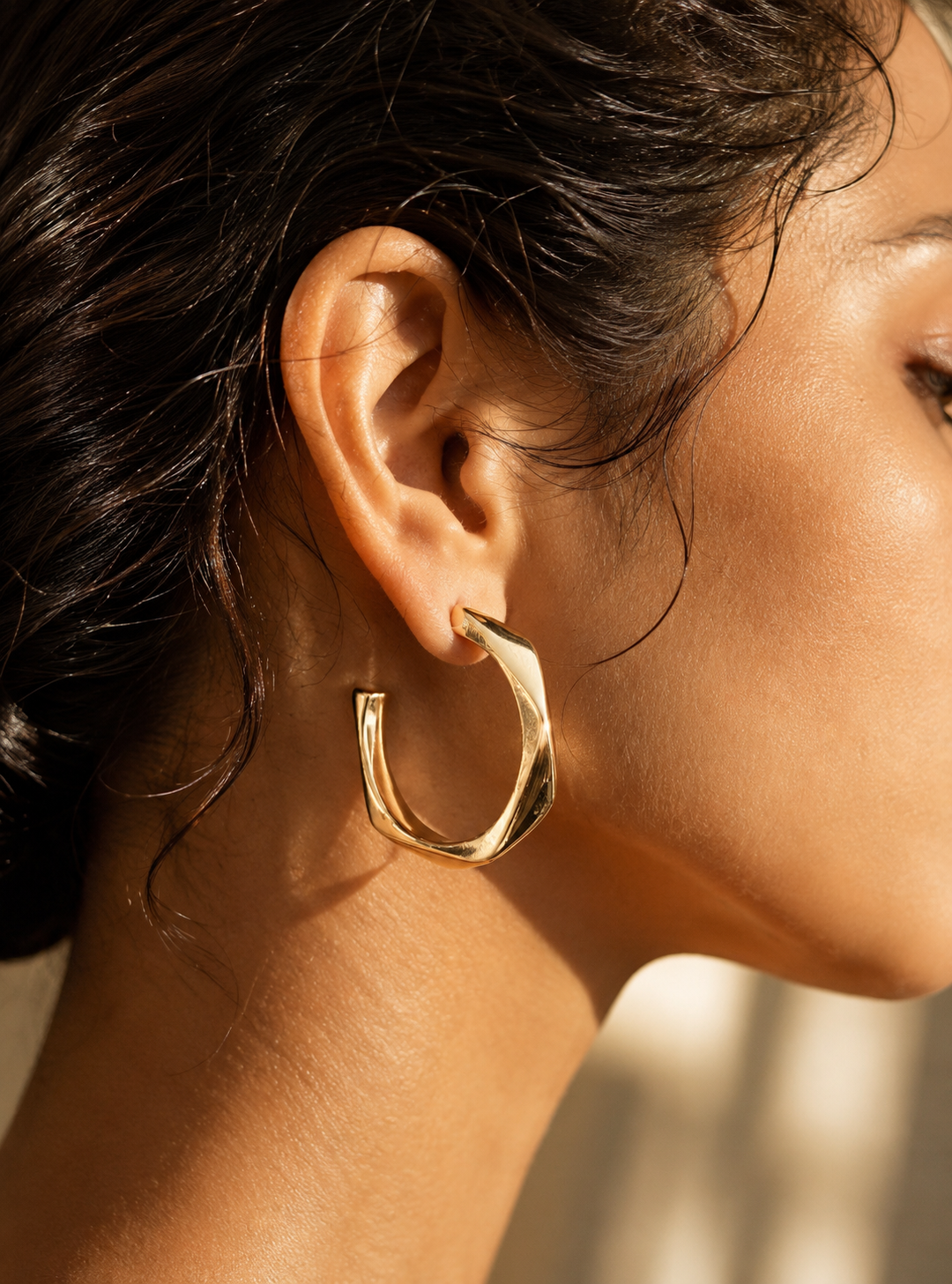 A flat-lay product photograph of a pair of oversized, elongated gold hoop earrings from the Long Twist collection, standing upright on porous white travertine stone steps under warm sunlight. The earrings feature a unique vertically stretched C-shape with an angular twisted design and a high-shine molten gold finish, fitted with clear plastic disc backings on the posts. The background is softly styled with delicate dried white flowers and a blurred Mediterranean architectural setting. As seen in GE011_long 