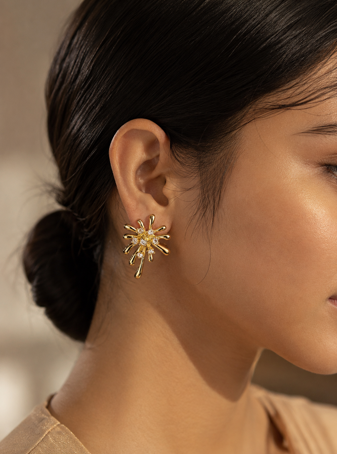 A flat-lay product photograph of a pair of oversized gold starburst stud earrings from the Diamond Pop collection. The earrings are displayed upright on porous cream-colored travertine stone blocks under warm, natural sunlight, showcasing a highly polished yellow-gold firework-like design adorned with scattered, sparkling clear crystals. The background features delicate dried white flowers and a soft, blurred view of a seaside architectural backdrop. As seen in GE012_Diamond POP_499.png.