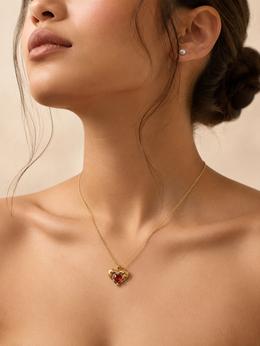 An elegant gold necklace with a red heart pendant and embedded crystals, displayed on a textured beige stone slab. The background features a minimalist, warm-toned aesthetic with cream ceramic vases, dried white hydrangea flowers, smooth gray river stones, and a soft white cloth, all illuminated by gentle sunlight and soft shadows.
