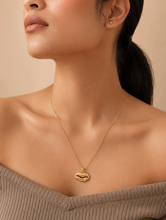 A gold chain necklace with a smooth, high-polish pendant shaped like a pair of lips, showcased on a white marble slab. The luxury lifestyle scene in GN002_gold lip.png is framed by shimmering, draped champagne satin fabric, a smooth beige river stone, fluffy dried bunny tail grass stems, and a sprig of green eucalyptus leaves under soft, warm lighting.