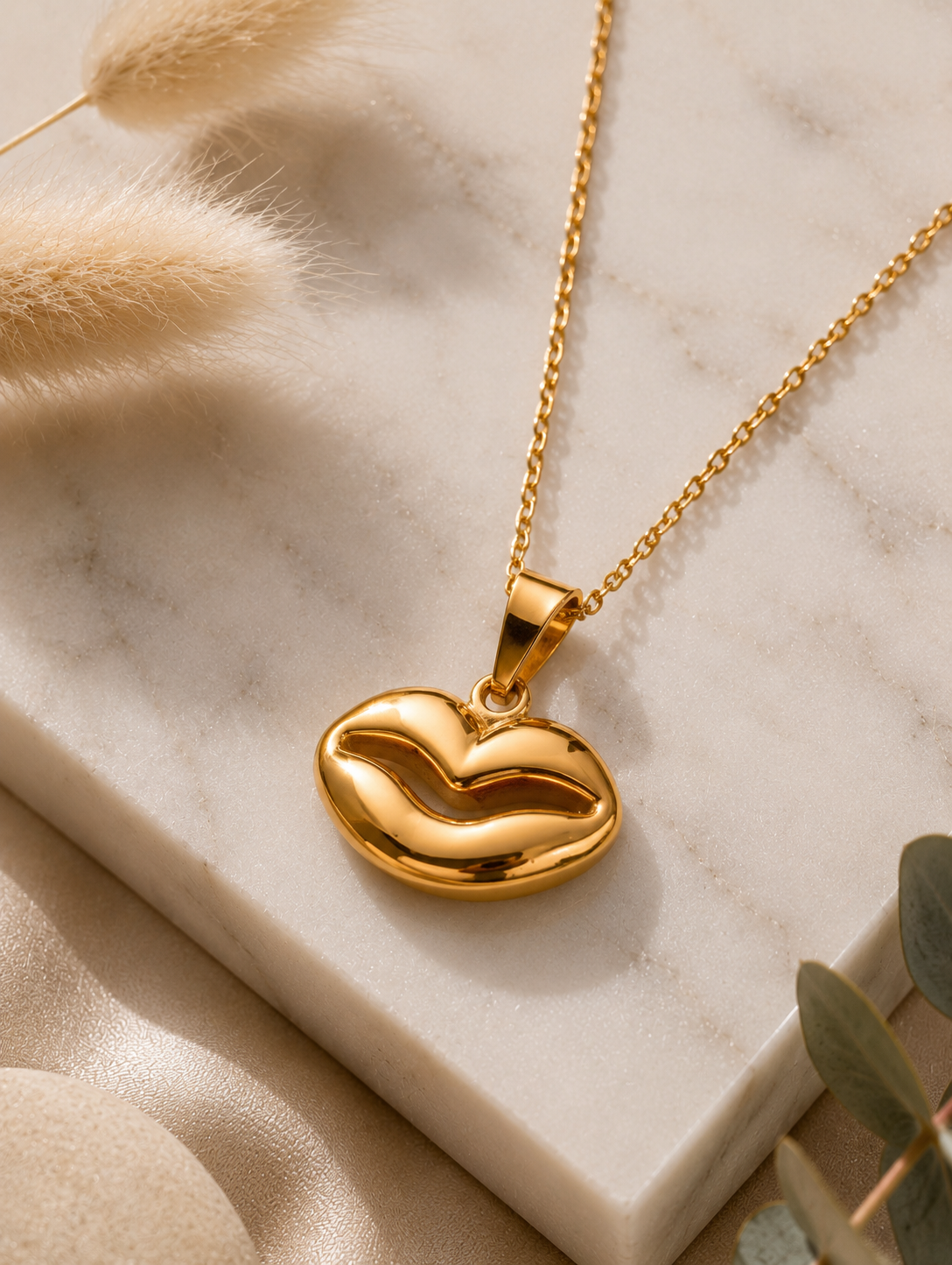A gold chain necklace with a smooth, high-polish pendant shaped like a pair of lips, showcased on a white marble slab. The luxury lifestyle scene in GN002_gold lip.png is framed by shimmering, draped champagne satin fabric, a smooth beige river stone, fluffy dried bunny tail grass stems, and a sprig of green eucalyptus leaves under soft, warm lighting.