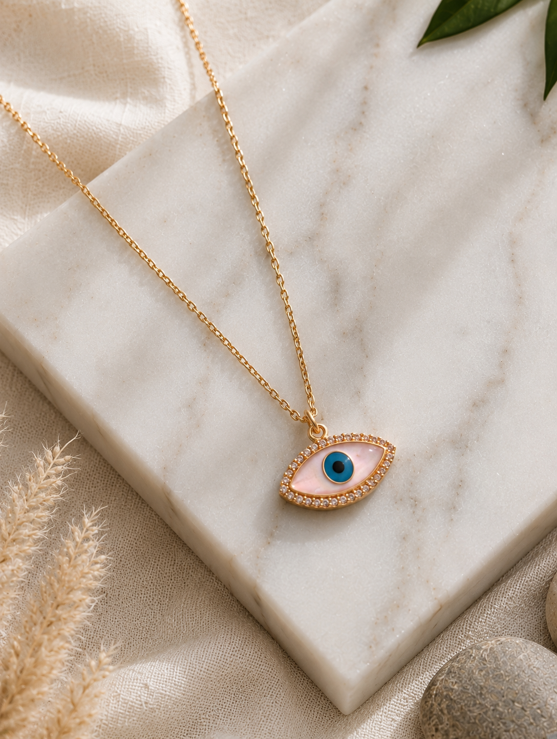 A gold chain necklace featuring an eye-shaped evil eye pendant with an iridescent pink mother-of-pearl inlay, a blue and black pupil, and a border of small sparkling crystals. The necklace in GN003_nazar.png is elegantly displayed on a square white marble block, surrounded by a textured linen cloth, dried pampas grass, smooth river stones, and a green leaf tip under warm, natural lighting.